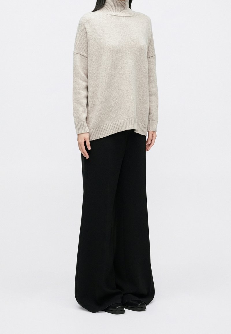 Personne portant un pull à col roulé beige avec des manches longues et un pantalon noir large et ample, debout devant un fond blanc uni.