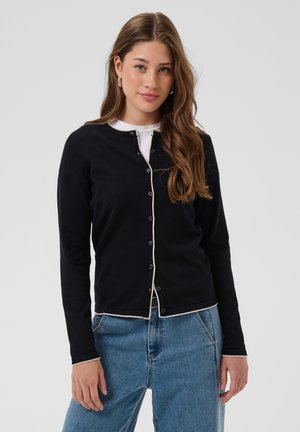 Jeune femme aux longs cheveux bruns portant un cardigan noir boutonné sur une chemise blanche et un jean bleu, debout devant un fond uni.