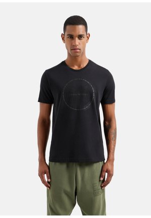 Camiseta de algodón negra con mangas cortas y cuello redondo. Presenta un logo circular texturizado con "EA7 EMPORIO ARMANI MILANO" en el centro.