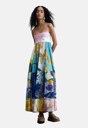 Žena sa kovrčavom kosom nosi šarenu cvjetnu maksi haljinu s tankim naramenicama i velike zlatne naušnice u obliku cvijeta, stoji s rukama iza leđa.