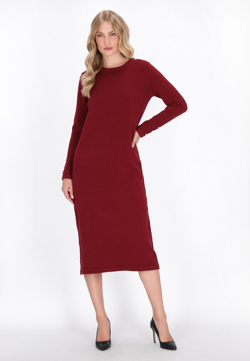 Burgunderfarbendes langärmliges Kleid mit schlanker Passform, Rundhalsausschnitt und Seitenschlitzen; mit Knopfdetails an den Ärmeln. Getragen mit schwarzen High Heels.