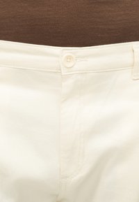 Close-up van de taille van gebroken-witte broek met knoopsluiting tegen een bruine stofachtergrond.