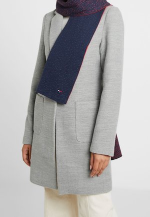 Femme portant un manteau gris mi-long avec des poches appliquées et une écharpe bleu marine à taches rouges avec un bord rouge.