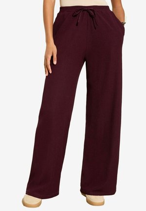 Pantalon en maille bordeaux à jambes larges avec taille à cordon, porté avec des chaussures beiges, mains dans les poches et le long du corps.