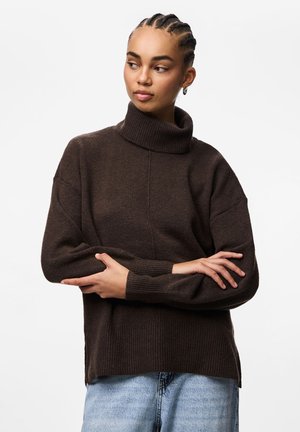 Femme aux cheveux tressés portant un pull à col roulé marron foncé et un jean bleu clair, les bras croisés, regardant vers sa gauche.