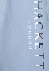 Un tessuto azzurro chiaro presenta un design testuale verticale con "HACKETT" e "LONDON" in una tipografia nera e bianca in contrasto. Texture morbida.