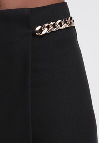 Černé kalhoty vysokého pasu s hladkým povrchem, v pase s akcentem zlatého řetězu, poskytující elegantní designový prvek.