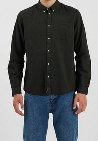 Homme portant une chemise noire à boutons avec une poche poitrine, associée à un jean en denim bleu, se tenant contre un fond uni.