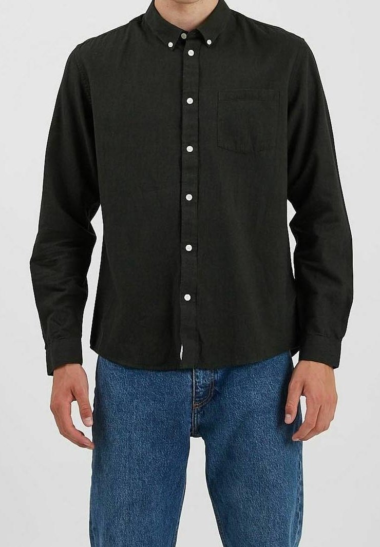 Homme portant une chemise noire à boutons avec une poche poitrine, associée à un jean en denim bleu, se tenant contre un fond uni.