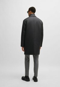 Cappotto imbottito nero con colletto alto, texture liscia e taglio dritto, indossato sopra pantaloni grigi a motivi e stivali neri.
