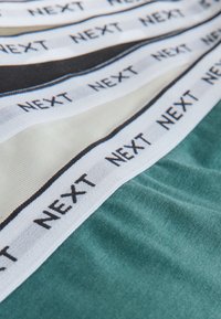 Gros plan de bandes de tissu avec le texte répété « NEXT » sur les bords blancs cousus sur des couches de tissu noires, blanches et vertes.