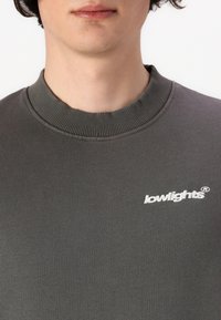 Tamsiai pilkas džemperis su raukinėta apykakle. Priekyje yra balta siuvinėta logo "lowlights". Minkšta medžiagos tekstūra.