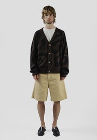 Braun gemusterter Cardigan mit V-Ausschnitt, großen Knöpfen und langen Ärmeln, kombiniert mit beigen Shorts und schwarzen Loafers. Minimalistischer Hintergrund.