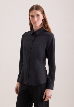 Kobieta o długich brązowych włosach, ubrana w dopasowaną czarną koszulę zapinaną na guziki i czarne spodnie, stojąca na jasnym, jednolitym tle.