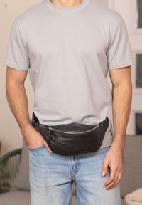 Marsupio in pelle nera con design curvo e dettagli in cerniera argentata, indossato sopra una maglietta grigio chiaro e jeans azzurri.