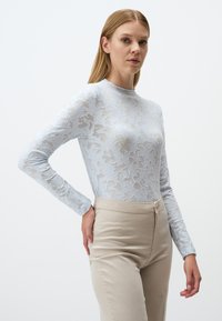 Top lungo a maniche lunghe di pizzo blu chiaro con motivi floreali, colletto alto, abbinato a pantaloni aderenti beige con zip frontale e leggera lucentezza.