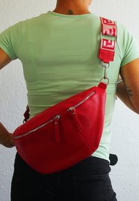 Roter Leder-Hüftbeutel mit zwei Reißverschlusstaschen, verstellbarem Gurt mit Logo und glatter Textur. Quer über den Körper getragen.