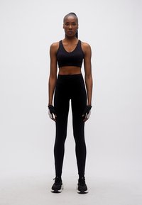 Αθλητική γυναίκα με μαύρο αθλητικό σουτιέν, κολάν με ψηλή μέση, γάντια και αθλητικά παπούτσια, στέκεται μπροστά σε απλό φόντο.