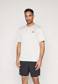 Camiseta deportiva blanca de manga corta y cuello redondo; cuenta con un pequeño logo negro en el pecho izquierdo. Combinada con pantalones cortos negros.