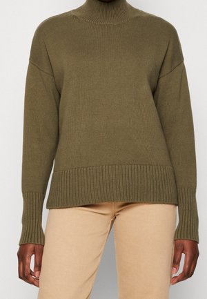 Person trägt einen olivgrünen gerippten Rollkragenpullover und beige Hose, steht vor einem schlichten hellen Hintergrund.