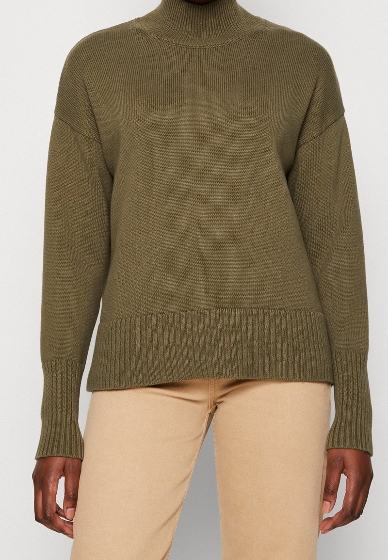 Personne portant un pull à col roulé côtelé vert olive et un pantalon beige, debout devant un fond clair uni.
