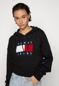 Crna pletiva kapuljača s rebrastom teksturom, špagom i geometrijskom logotipom u crvenoj, bijeloj i plavoj boji na prednjem dijelu.