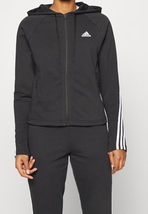 Kvinde iført en sort Adidas hættetrøje med lynlås og hvide striber på ærmerne samt matchende sorte bukser mod en ensfarvet baggrund.