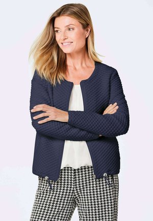 Veste matelassée bleu marine avec un col rond et des poches zippées, associée à un pantalon à motifs pied-de-poule noir et blanc. Texture de tissu lisse.