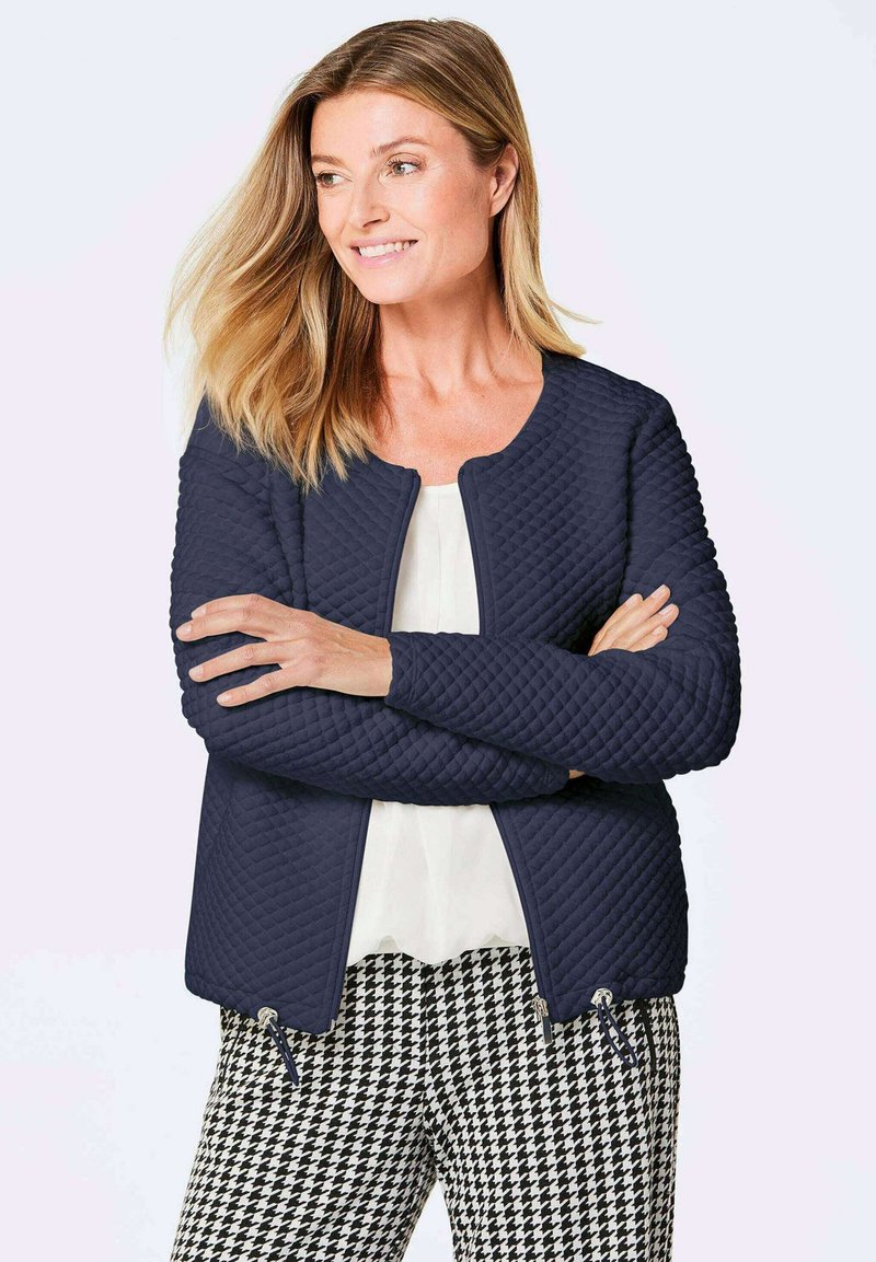 Gesteppten navy Jacke mit rundem Ausschnitt und Reißverschlusstaschen, kombiniert mit schwarzer und weißer Hahnentritt-Hose. Glatte Stoffstruktur.