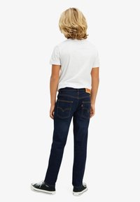Jeans denim w ciemnoniebieskim kolorze, slim fit, z złotymi szwami i charakterystycznymi tylnymi kieszeniami. Połączone z jednolitą białą koszulką i czarnymi sneakersami.