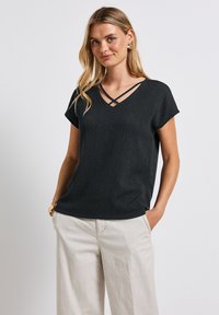 Černý texturovaný top s krátkým rukávem a výstřihem do V, která má křížené ramínka. Stylizováno s světle zbarvenými, volně střiženými kalhotami pro kontrast.