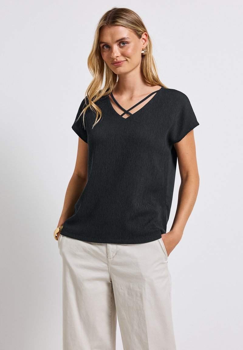 Černý texturovaný top s krátkým rukávem a výstřihem do V, která má křížené ramínka. Stylizováno s světle zbarvenými, volně střiženými kalhotami pro kontrast.