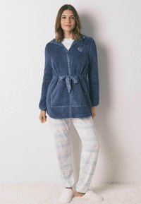 Laivastonsininen fleece-vetoketjullinen takki, jossa on kiristysnauha vyötäröllä ja kaksi etutaskua, yhdistettynä valkoisiin kuvioituihin pyjamahousuihin ja pehmeisiin tohveleihin.