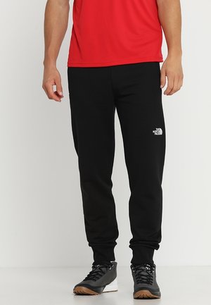 Persoon draagt zwarte The North Face trainingsbroek, rood shirt en zwarte sportschoenen met witte zolen op een effen achtergrond.