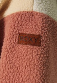Chaqueta de forro polar en colores rosa y crema con un acabado texturizado. Presenta una etiqueta de cuero marrón con "ROXY" grabado en ella.