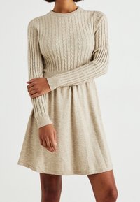 Abito in maglia a trecce beige a maniche lunghe con vita aderente e gonna svasata, indossato da una persona con le braccia incrociate contro uno sfondo neutro.