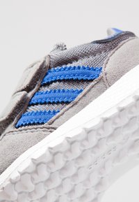 Harmaa urheilukenkä sinisillä raidallisilla yksityiskohdilla ja teksturoidulla kangaspäällisellä. Kumipohjassa on mehiläiskennoa muistuttava kuvio, joka parantaa pitoa.