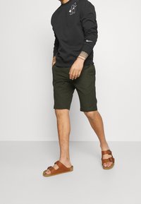 Sudadera negra con logo blanco, pantalones cortos verde oliva y sandalias marrones con correas dobles. Corte recto; conjunto informal.