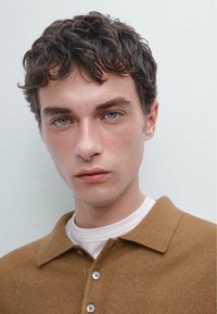 Hnědý pletený polo shirt s límečkem a detailem knoflíku, vrstvený přes bílý tričko. Model má krátké, kudrnaté tmavé vlasy a modré oči.