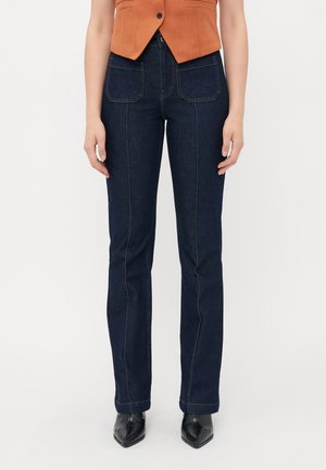 Kvinne iført mørkeblå rette jeans med frontlommer, oransje ermeløs knappetopp og svarte spisse støvletter.