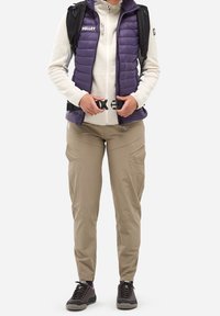 Person, der beige Hosen trägt, eine lila Steppweste über einem cremefarbenen Reißverschluss-Pullover hat und den Schnalle des schwarzen Rucksacks schließt. Steht vor weißem Hintergrund.