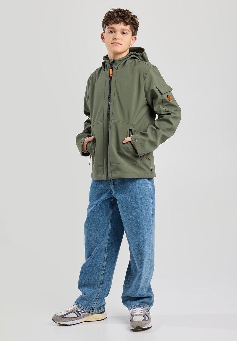 Olive green softshell jacket with zip closure, adjustable hood, two front pockets, and patch detail on the left sleeve. Worn with light blue jeans.
