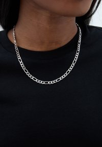 Sidabrinė grandinėlė su sujungtomis grandinėlėmis, dėvima virš juodos apvalios iškirptės marškinėlių, demonstruojanti lygų metalinį paviršių ir glotnų tekstūrą.