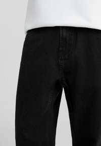Jeans de mezclilla negra con un corte clásico, textura sutil y costuras definidas. Se ve que se combinan con una camisa blanca en la parte superior.