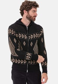 Schwarz-beiger, gemusterter Reißverschluss-Pullover mit geometrischen Designs, gerippten Bündchen und Saum. Verfügt über einen hohen Kragen und ein strukturiertes Strickmaterial.