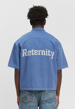 Blaue Denim-Hemd mit kurzen Ärmeln und dem Wort "Reternity" in großen weißen Buchstaben auf dem Rücken. Mit lockerer Passform und Kragen.