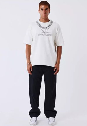 Junger Mann mit kurzen rötlichen Haaren, der ein weißes oversized T-Shirt mit Silberketten-Print, schwarze Hose, weiße Sneaker und silbernen Schmuck trägt.