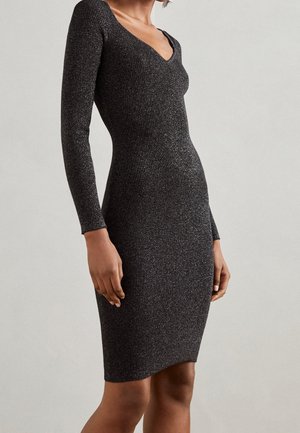 Kobieta nosząca dopasowaną, czarną błyszczącą dzianinową sukienkę z długim rękawem, sięgającą do kolan, z dekoltem w serek, na jednolitym jasnym tle.