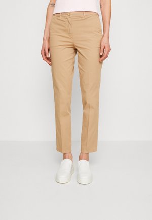 Femme portant un pantalon beige à jambes droites et des chaussures blanches sans lacets, debout devant un fond clair uni.