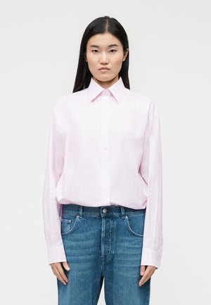 Pitkähiuksinen nainen, jolla on päällään vaaleanpunainen napitettava paita ja korkeavyötäröiset siniset farkut, seisoo yksinkertaista valkoista taustaa vasten.